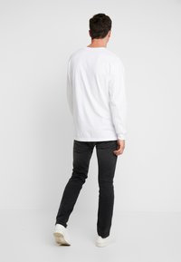 Camiseta blanca de manga larga, de material de algodón, corte relajado, combinada con jeans negros ajustados y zapatillas blancas, vista desde la parte de atrás.