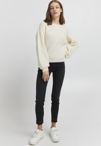 Pull en tricot crème avec un motif texturé, manches larges, associé à un jean noir ajusté et des baskets blanches. Ensemble simple et décontracté.