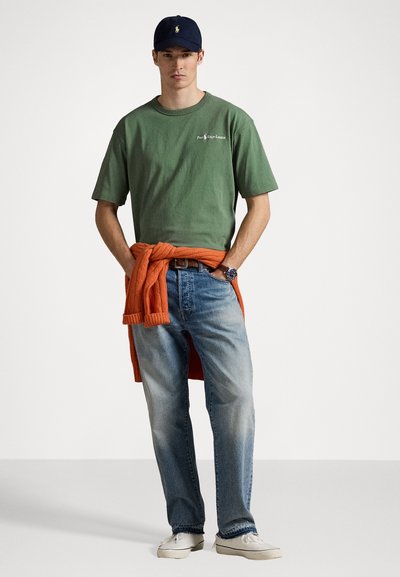 Green cotton t-shirt with logo, light blue denim jeans, wrapped orange knit sweater, and white sneakers. Navy cap with logo accent.