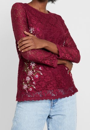 Woman wearing a long-sleeve burgundy lace top with floral embroidery on the side, paired with light blue denim jeans, arms crossed.