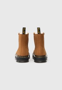 Botine din piele maro cu talpă din cauciuc negru, cusături galbene și bucle de prindere cu textul "CU TALPĂ ELASTICA". Finisaj texturat.