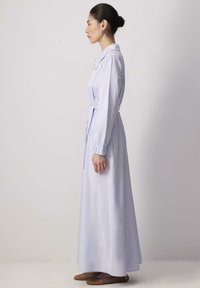 Robe longue bleu clair avec un col, des manches longues et une ceinture à nouer à la taille. Tissu lisse avec un léger éclat. Portée avec des chaussures plates marron.