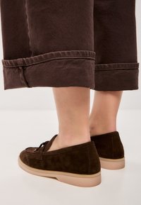 Bruine suede instappers met een slanke beige rubberen zool, voorzien van een vetersluiting en een ronde neus, naast opgerolde bruine broeken gepresenteerd.