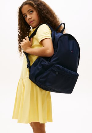 Dziewczynka z kręconymi włosami, ubrana w żółtą sukienkę i niosąca dużą granatową plecak na jednym ramieniu, patrząca nieco na bok.