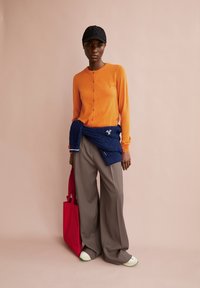 Orange long-sleeve cardigan, navy sweater tied at waist, loose gray trousers, black cap, red tote bag, white and green sneakers.