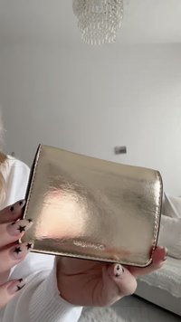 Portafoglio metallico dorato con una texture liscia, forma rettangolare e bordi cuciti. Presenta il nome del marchio in rilievo in basso.