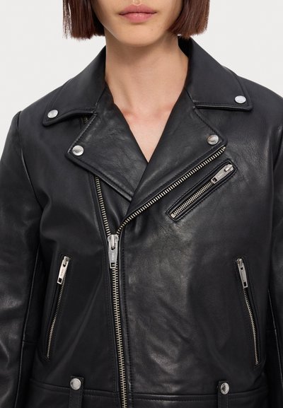 Femme portant une veste de motard en cuir noir avec des fermetures éclair argentées et des boutons-pression, fermeture éclair centrale avant partiellement ouverte.