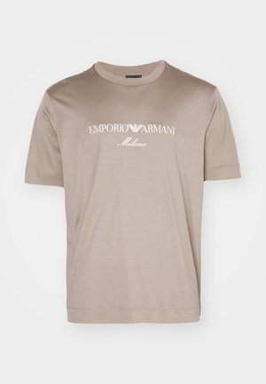 Beige színű, rövid ujjú póló, puha anyagból készült, amelyen fehér színű "EMPORIO ARMANI" és "Milano" logó látható a mellkason.