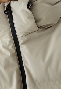 Beige gewatteerd jack met een zwarte rits. Het materiaal oogt glad en lichtgewicht, met een hoge kraag en minimale stikdetails.