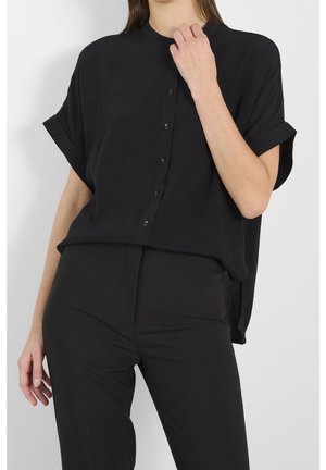 Mujer con una camisa negra de manga corta, holgada, abotonada, con los puños remangados y pantalones negros ajustados sobre un fondo liso.