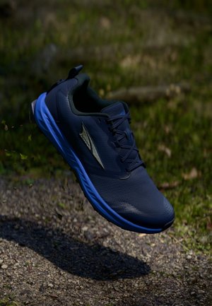 Tmavě modrá běžecká bota se světle modrou podrážkou se objevuje ve vzduchu nad štěrkovou cestou s trávnatým pozadím.