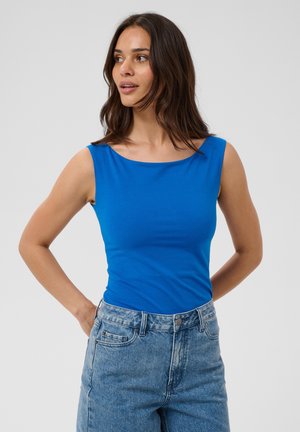 Vrouw met schouderlang donker haar, draagt een felblauw mouwloos topje en lichtblauwe hooggetailleerde jeans, kijkt opzij met handen achter de rug.