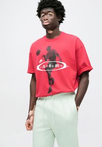 Κόκκινο βαμβακερό μπλουζάκι με μαύρο σχέδιο καλαθοσφαιριστή, με λευκό λογότυπο Nike και κείμενο «Air Jordan». Συνδυάζεται με ανοιχτό πράσινο παντελόνι.