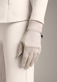 Beżowe rękawiczki wykonane z gładkiego materiału z dopasowanym mankietem, ozdobione małym czarnym elementem metalowym oraz czterema szwami biegnącymi przez dłoń.