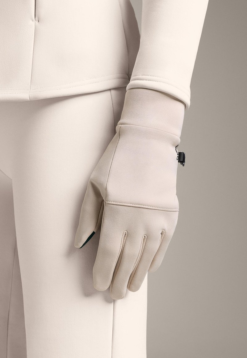 Béžové rukavice z hladkého materiálu s přiléhavým zápěstím, vybavené malým černým kovovým detailem a čtyřmi švy přes dlaň.