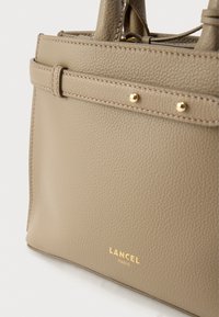 Beežist nahast käekott tekstuurse pinnaga, millel on õhuke disain, kuldse tooniga metalldetailid ja logo põhjas embosseeritud.