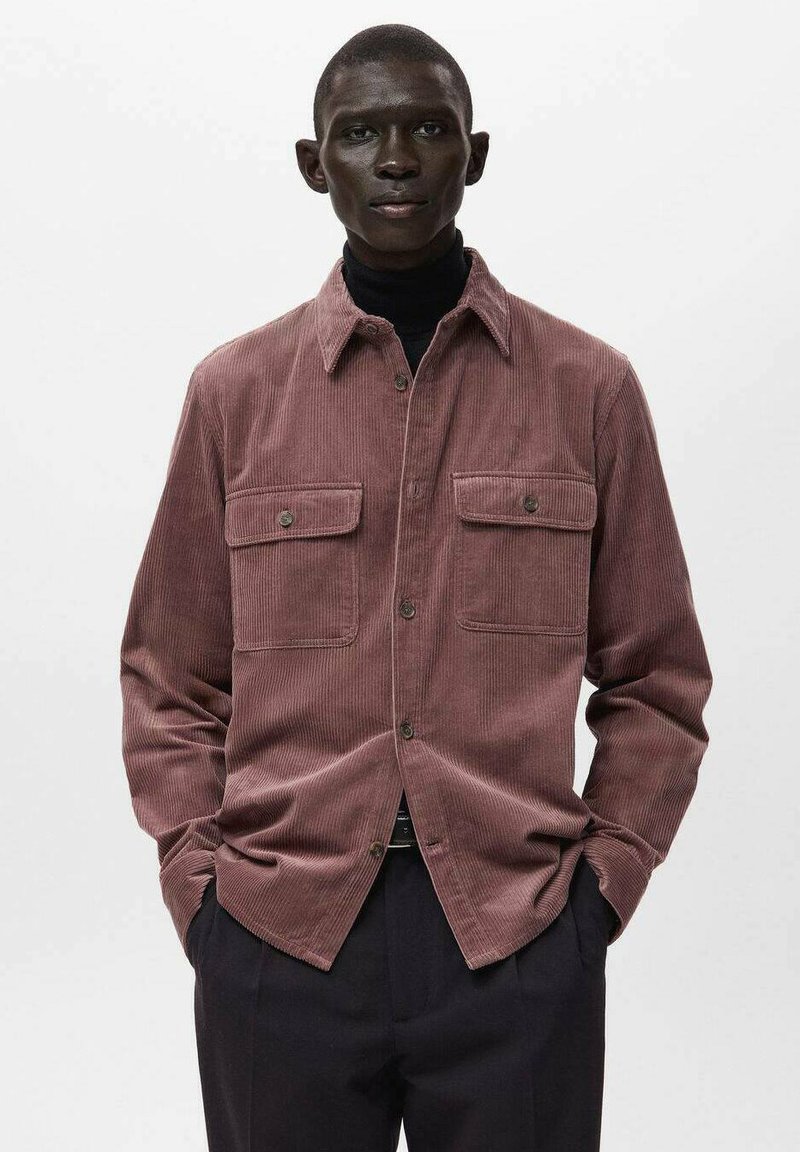 Manchester-skjorta i mauve med en texturerad yta, med två bröstfickor, knäppning och skjortkrage. Bärs över en svart polotröja.
