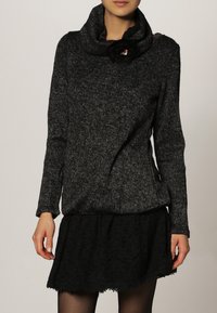 Svart, strukturerad tröja med halsringning och en blommig accent. Bärs över en svart, lager-på-lager kjol med ett spetsmönster. Långa ärmar.