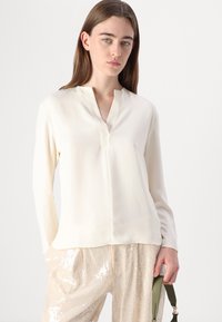 Blusa a maniche lunghe color crema con scollatura a V e leggera lucentezza. Abbinata a pantaloni beige chiaro con paillettes, creando un contrasto di texture uniforme.