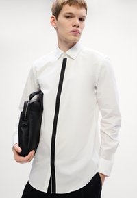 Witte button-down shirt met een centrale zwarte gestructureerde streep, lange mouwen en een gebogen zoom. Een zwarte leren tas wordt onder één arm vastgehouden.