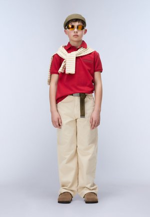 Boy wearing red polo, beige pants, striped sweater over shoulders, brown shoes, green cap, and reflective sunglasses standing against plain background.