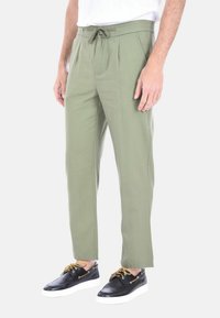 Pantaloni verde oliva a sigaretta con pieghe, cintura con cordino e tasche laterali; abbinati a scarpe nere con suole bianche e lacci marroni.
