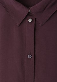 Chemise habillée violet foncé avec un col pointu, un bouton métallique en haut et un bouton en plastique en bas cousus sur la patte de boutonnage avant.