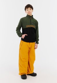 Fleecejacke in Grün und Schwarz mit orangefarbenem Logo, halber Reißverschluss vorne und seitlicher Tasche. Kombiniert mit lockeren gelben Hosen und schwarzen Stiefeln.