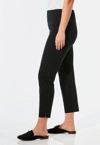 Pantalon noir extensible avec une taille élastique, coupe raccourcie, fentes latérales à l'ourlet, associé à des chaussures noires à enfiler, design minimaliste.
