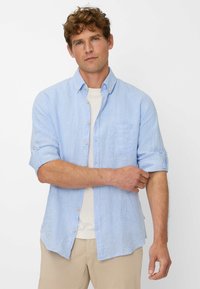 Camicia azzurro chiaro con bottoni, realizzata in un tessuto texture. Presenta una tasca sul petto e maniche arrotolate. Indossata sopra una maglietta bianca.