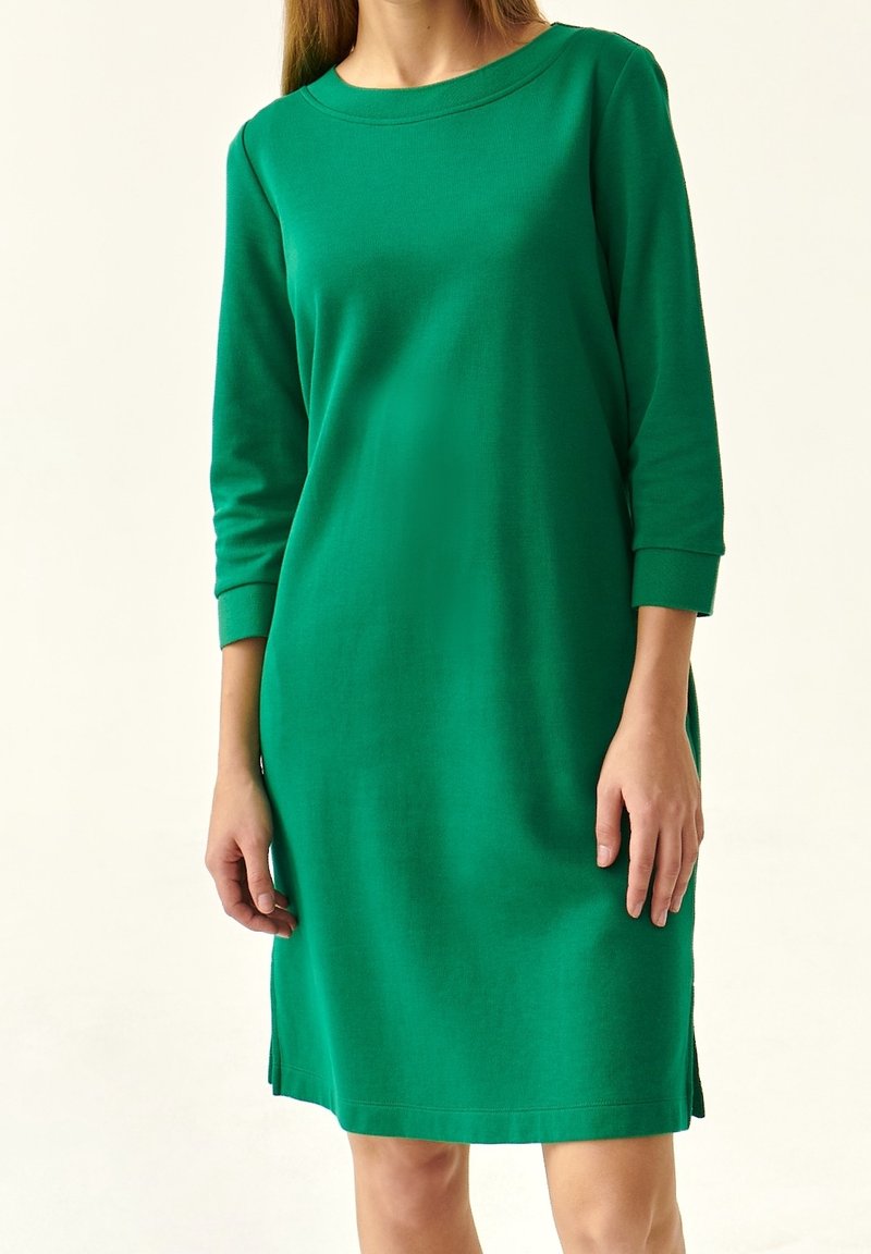 Femme portant une robe verte longueur genou avec des manches trois-quarts, debout devant un fond clair uni.