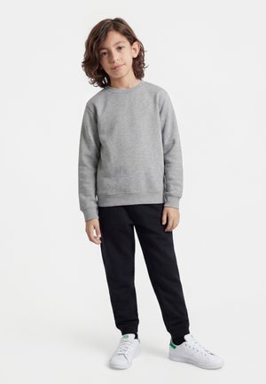 Niño con cabello castaño hasta los hombros, vistiendo una sudadera gris, pantalones negros y zapatillas blancas, de pie sobre un fondo blanco.