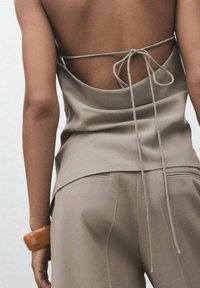 Haut en satin beige avec un dos ouvert, noué avec un fin cordon. Le modèle porte un pantalon beige assorti avec des poches latérales et un accessoire en bois.