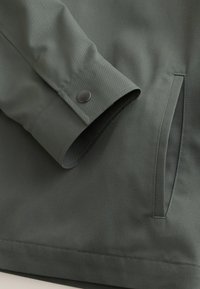 Manche de veste en tissu vert avec un poignet à bouton-pression métallique et une poche latérale verticale cousue sur le corps.