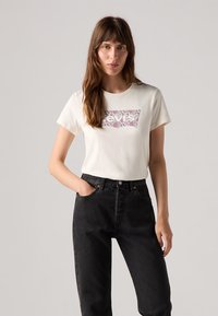 Cremfarbene Baumwoll-T-Shirt mit einem bunten floralen Grafik-Logo auf der Vorderseite, kombiniert mit hoch taillierten schwarzen Denim-Jeans.