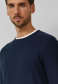 Bebaarde man met lichte huid en blauwe ogen, gekleed in een marineblauwe trui met ronde hals over een wit ondershirt, tegen een effen grijze achtergrond.