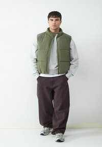 Eine plüschige grüne Weste über einem grauen Sweatshirt, kombiniert mit lockeren braunen Hosen und grauen Sneakers. Die Weste hat einen hohen Kragen und einen Reißverschluss.