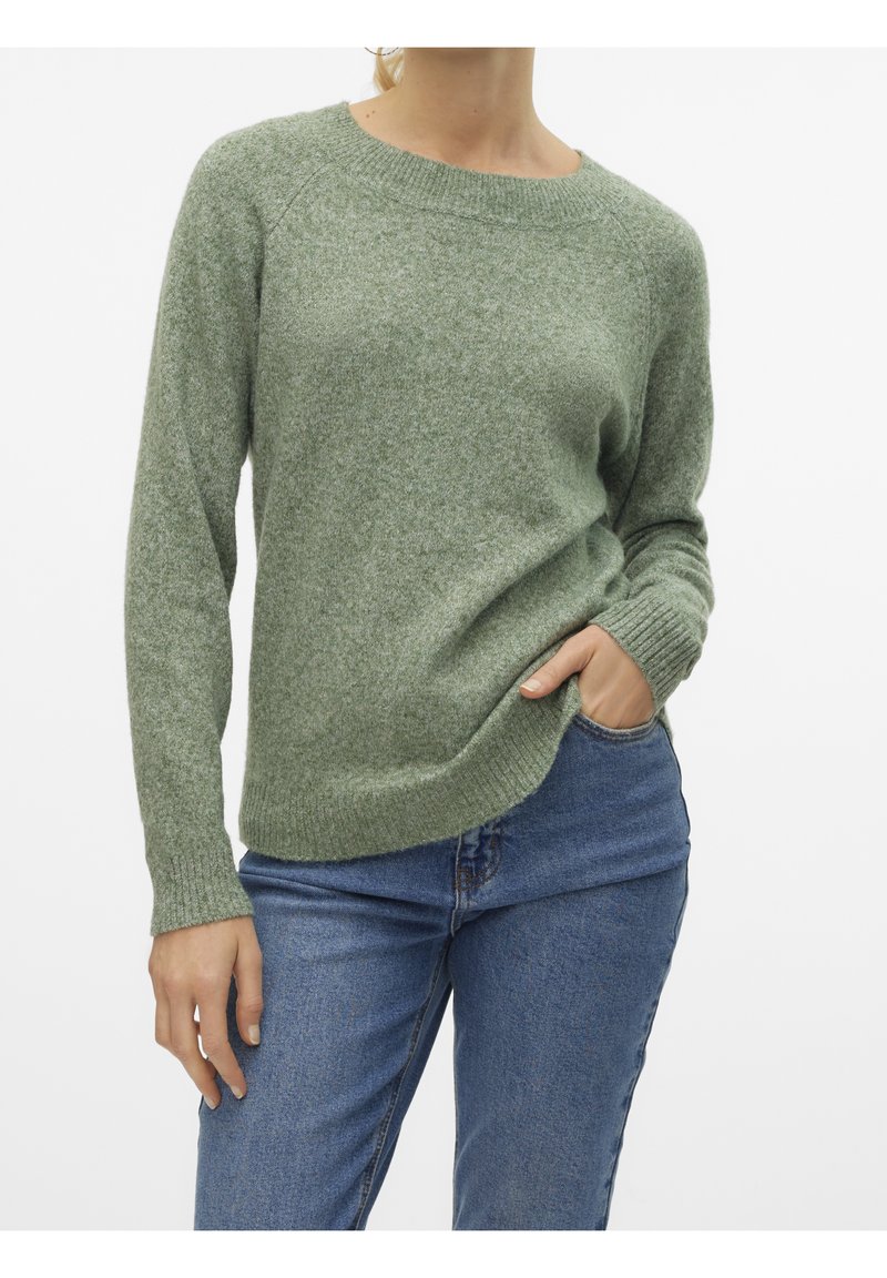 Femme portant un pull en maille vert clair avec poignets et ourlet côtelés, associé à un jean en denim bleu, main dans la poche, sur fond blanc.