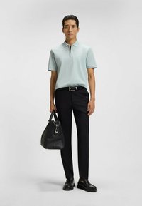 Ljusblå polo med dragkedja, svarta byxor och en texturerad svart handväska med handtag. Skorna är svarta slip-on skor.