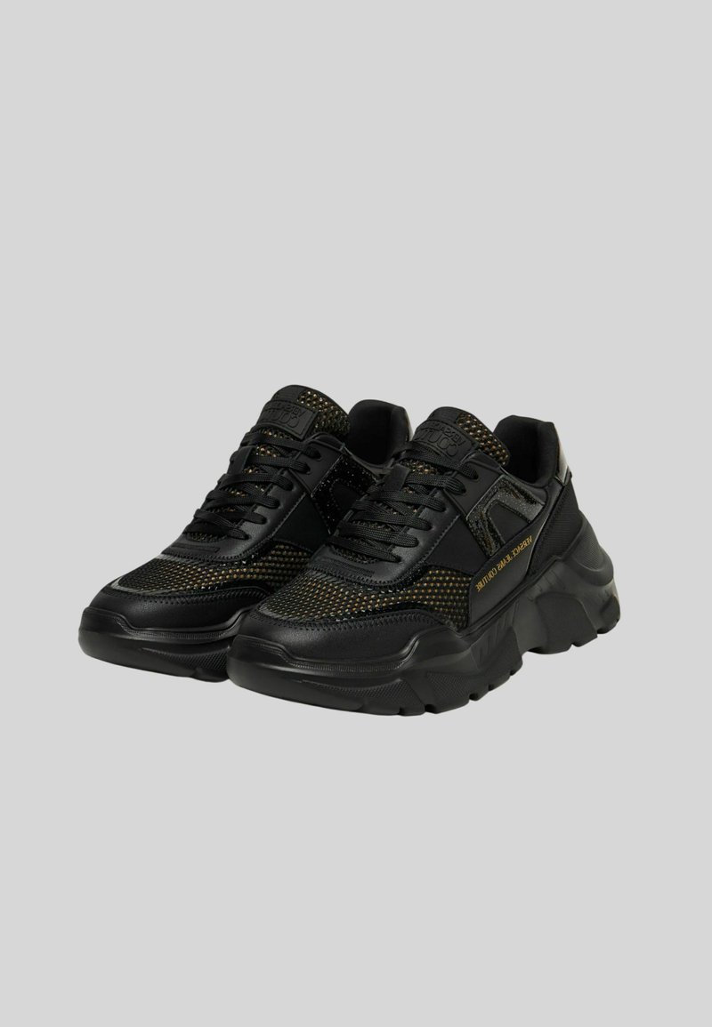 Baskets noires avec des détails en mesh et une semelle épaisse. Elles présentent des lacets noirs, des accents texturés et un logo doré sur le côté.
