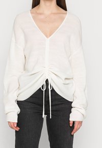 Weißer Rippstrickpullover mit tiefem V-Ausschnitt, gerafftem Design an der Taille und verstellbaren Drawstrings. Lange Ärmel und weiche Textur.