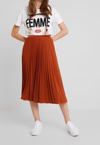 Camiseta blanca con gráfico negro "FEMME", combinada con una falda midi plisada naranja y zapatillas blancas. Textura suave, ajuste casual.