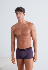 Boxer da uomo viola con vita nera caratterizzata da testo rosso "HUGO". Il materiale sembra liscio con un design aderente. Il modello ha un fisico tonico.
