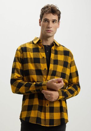 Camicia di flanella a quadri gialli e neri con colletto a bottoni, maniche lunghe e tasca sul petto. Texture morbida con un motivo prominente.