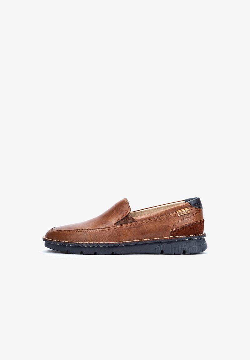 Zapato marrón de cuero tipo mocasín con punta redonda, paneles laterales elásticos y un talón de un azul oscuro contrastante, que presenta detalles de costura visibles.