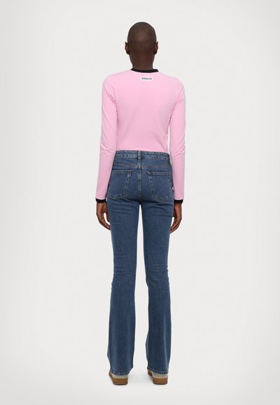 Light pink long-sleeve top with a black neckline, worn with blue flared jeans. Features a logo at the back and a fitted design.