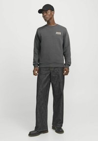 Grå sweatshirt med trykt "AWAKEN" logo, parret med mørke vide jeans og sorte støvler. Modellen bærer en sort kasket.