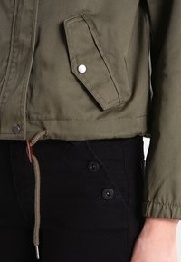 Olijfgroene canvas jas met een borstzak met knopen, trekkoordzoom en zilverkleurige details. Gecombineerd met zwarte broeken met knoppenaccenten.