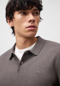 Polo grigia con un tessuto a maglia testurizzato, caratterizzata da un'apertura con tre bottoni e un piccolo logo. Indossata sopra una t-shirt bianca a girocollo.