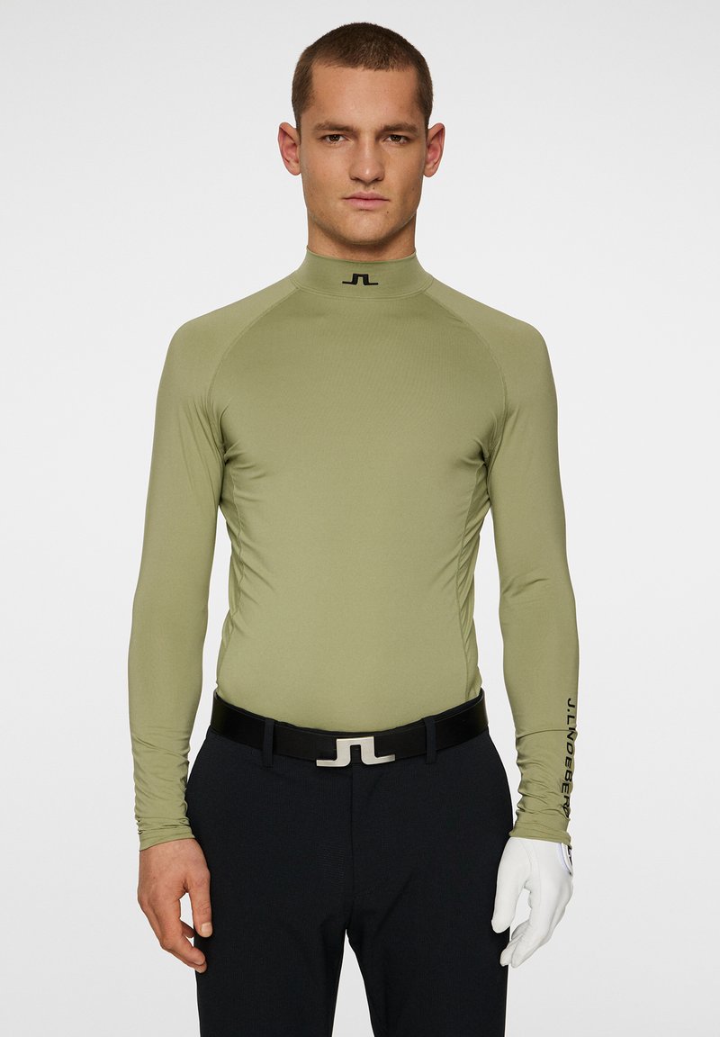 Långärmad, olivgrön golftröja med hög krage och logotyp vid halsen. Stretchig, åtsittande design; kombinerad med mörka byxor och ett svart bälte.